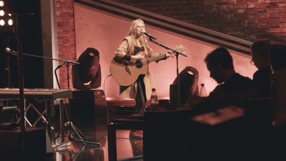Eine junge Frau mit einer Gitarre steht auf einer Bühne und singt in ein Mikrofon  