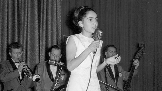 Die Sängerin Paola steht 1964 auf einer Bühne und singt. Hinter ihr die Begleitband. © Paola 