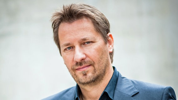 Wolfgang Norman Dalheimer steht mit dunkelblauem Hemd und Jacket vor einem grauen Hintergrund. © Steffen Z. Wolff 