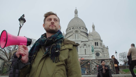 Moderator Consi mit einem Megafon vor der Basilika Sacré-Cœur in Paris. © NDR Foto: Screenshot