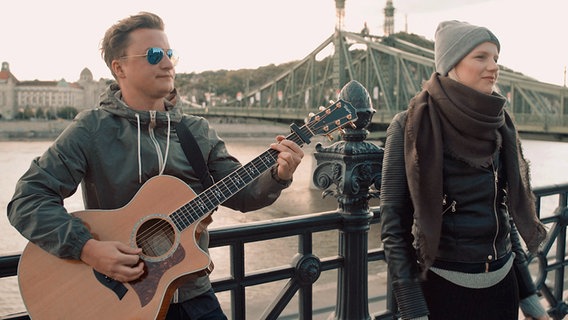 Gitarrist David Drink und ESC-Kandidatin Levina stehen in Budapest vor der Freiheitsbrücke. © NDR 