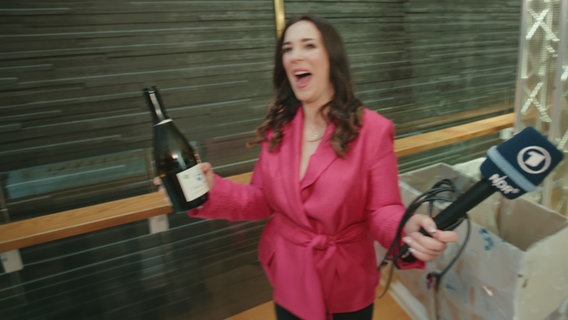 Alina Stiegler mit einer Flasche Sekt und einem NDR Mikrofon in der Hand. © NDR Foto: Screenshot