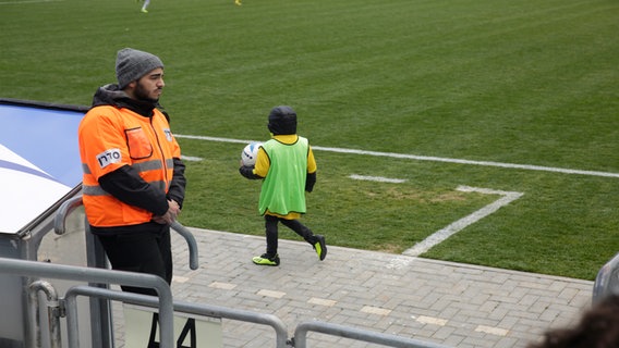 Ein kleines Ballkind, dass in einer dicken Jacke eingepackt einen Ball am Spielfeldrand in der Hand hält.  