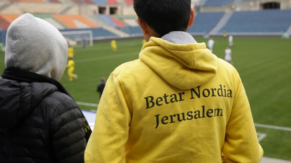 Ein Junge trägt einen gelben Hoodie mit der Aufschrift "Betar Nordia Jerusalem“ auf dem Rücken.  