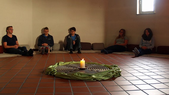 Fünf Jugendliche sitzen an Wände gelehnt auf Polstern und betrachten eine Kerze in der Mitte des Raumes © NDR Foto: Irving Wolther