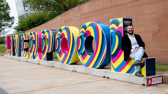 Irving Wolther sitzt auf einem großen Liverpool-Schriftzug in den diesjährigen ESC-Farben. © NDR Foto: Claudia Timmann