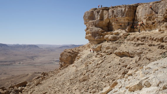 Ein riesiger Krater und eine Felswand, an der sich gesicherte Kletterer abseilen.  