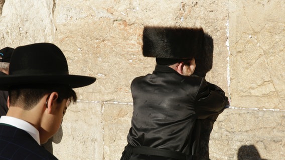 Ein Jude mit einem großen, mit Fell bezogenem Hut, lehnt an der Klagemauer und betet.  