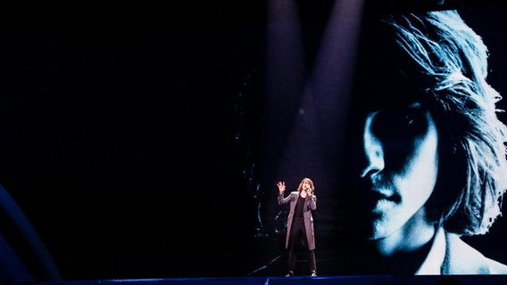 Isaiah steht auf der dunklen Bühne, sein Großporträt leuchtet blau im Hintergrund. © Eurovision.tv Foto: Thomas Hanses
