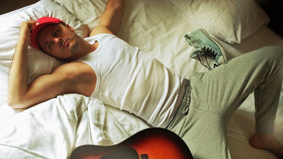 Daniel Kajmakoski liegt grübelnd mit rotem Basecap, weißem Unterhemd und Jogginghose auf einem Bett  Foto: Andreja Damjanovic