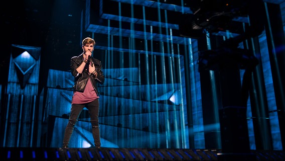 Justs steht vor blauen dunklen abstrakten Mustern auf dem Background-Monitor. © eurovision.tv Foto: Thomas Hanses (EBU)