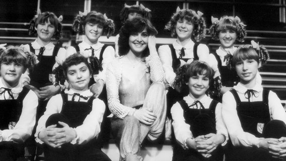 Die Sängerin Paola Felix sitzt 1980 in der ZDF Hitparade auf einer Treppe, umringt von Mädchen mit Zöpfen, die alle gleich angezogen sind. © Paola 