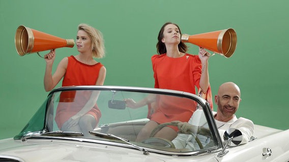 Der türkische Sänger Serhat fährt in einem weißen Auto, auf dem zwei Frauen mit orangenen Kleidern sitzen. © Klaus Roethliesberger 