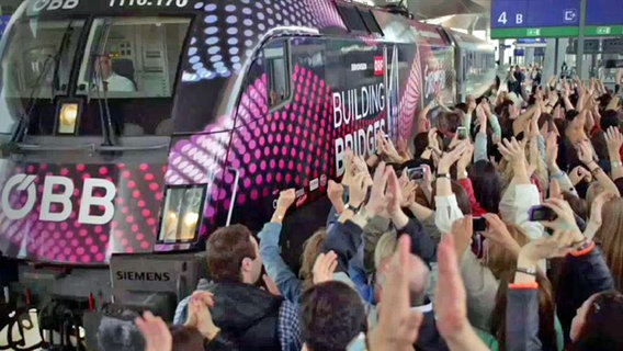 Ein Zug mit dem Logo des ESC 2015 "Building Bridges" fährt im Bahnhof ein und wird von Hunderten jubelnder Fans begrüßt. © ORF 